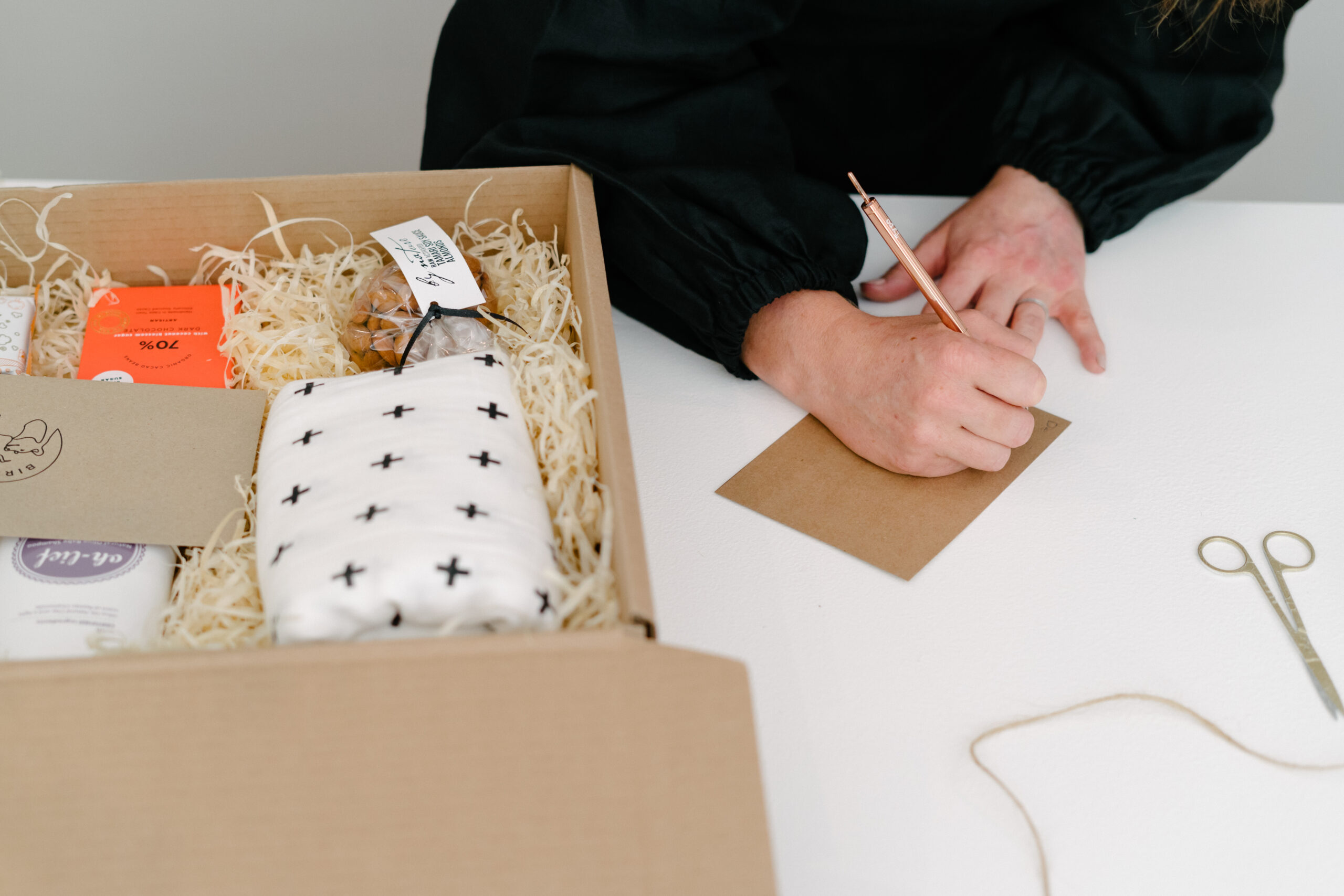 A shot of a packed gift box containing an assortment of items including a black and white muslin blanket, a slab of chocolate, a bag of nuts, a bottle of baby wash. A person can be seen writing a note on a brown card with a pair of scissors and some packing material on the table nearby